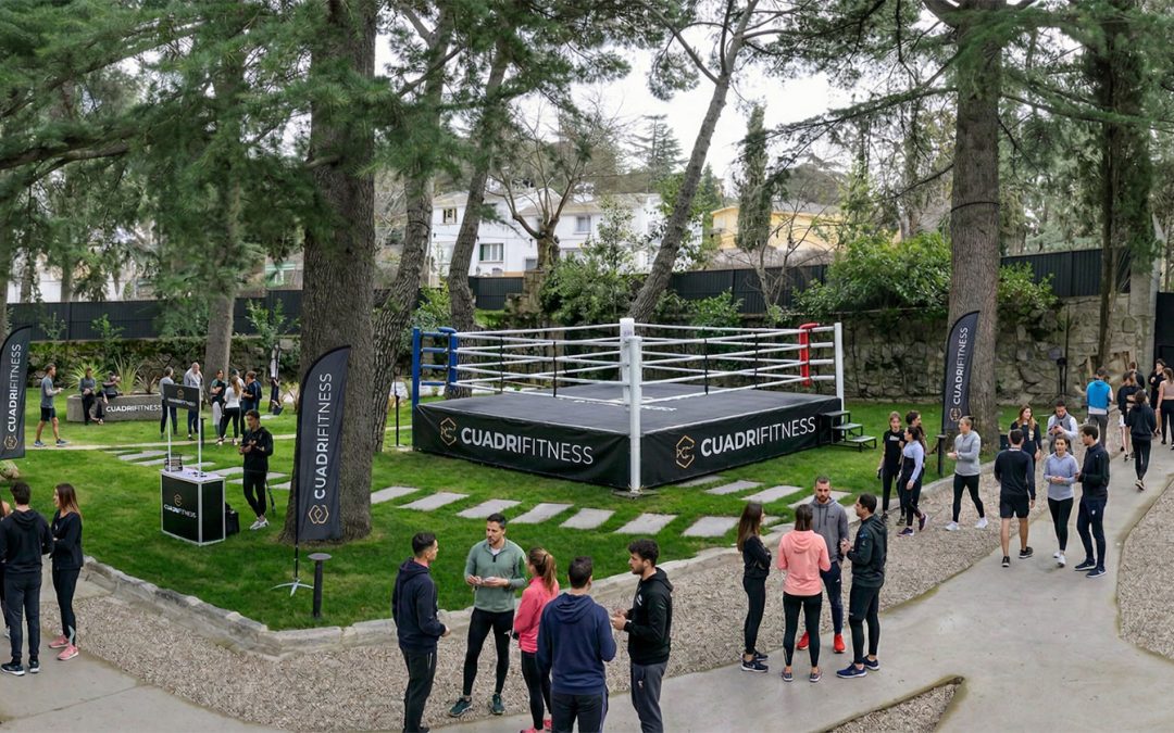 Un evento deportivo al aire libre en un jardín arbolado, con un ring de boxeo profesional de color negro en el centro. Grupos de personas con ropa deportiva conversan y caminan por senderos de grava alrededor de zonas de césped bien cuidadas.