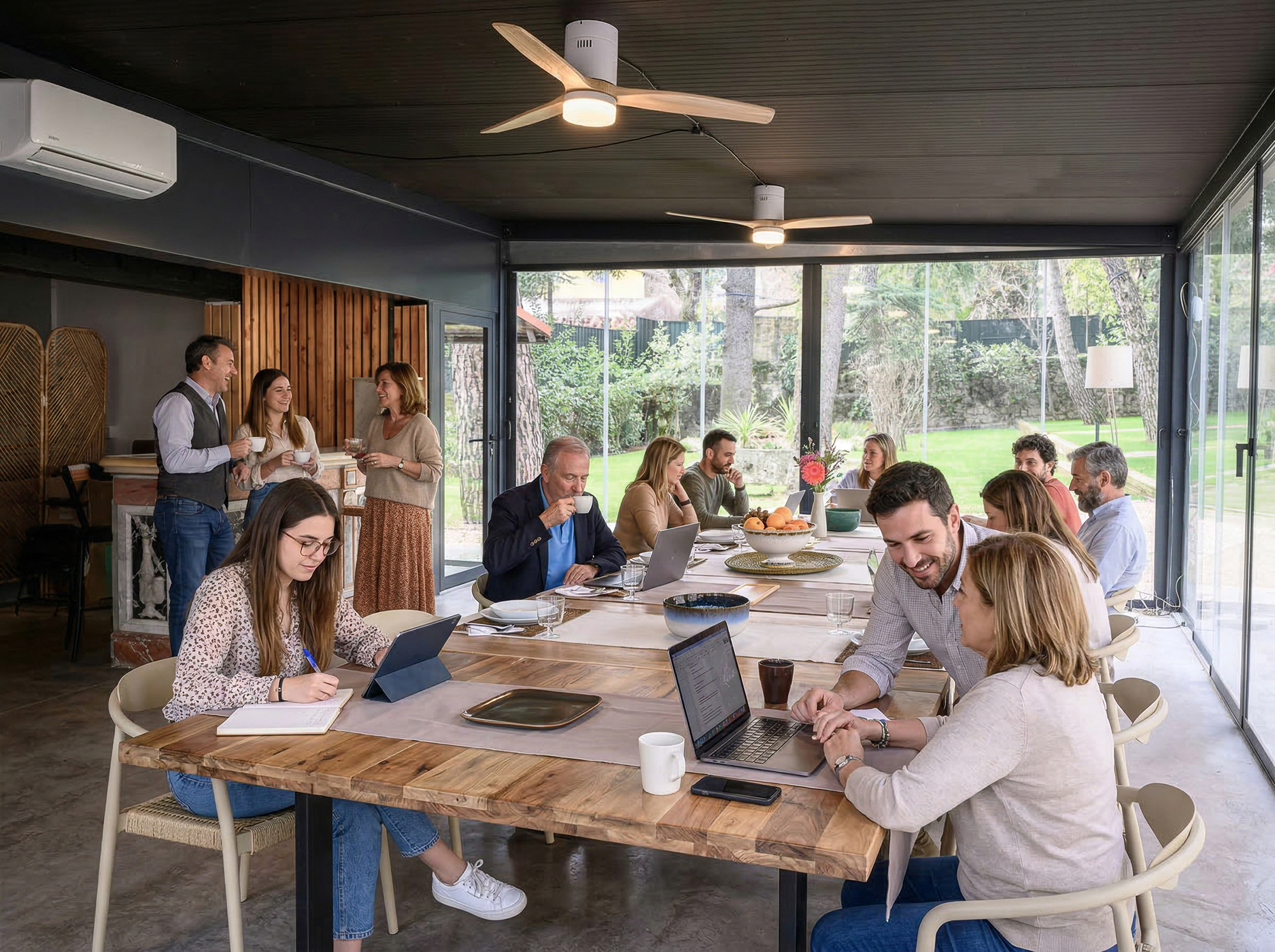 Un grupo de personas trabajando y colaborando en una sala de reuniones moderna y luminosa con grandes ventanales que dan a un jardín. Se ven mesas de madera, ordenadores portátiles, tabletas y personas conversando de pie al fondo.