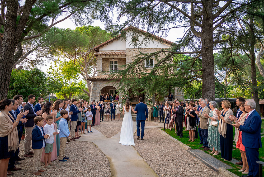 Una pareja de novios, de espaldas, camina por un sendero de grava hacia una casa de piedra y estuco rodeada de árboles. Están flanqueados por dos filas de invitados aplaudiendo en una ceremonia de boda al aire libre. La novia lleva un vestido blanco y el novio un traje azul. La luz del sol se filtra a través de las ramas.